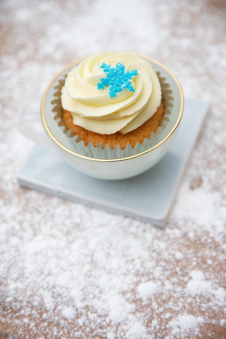A cupcake decorated with light frosting and a sugar snowflake in a cup