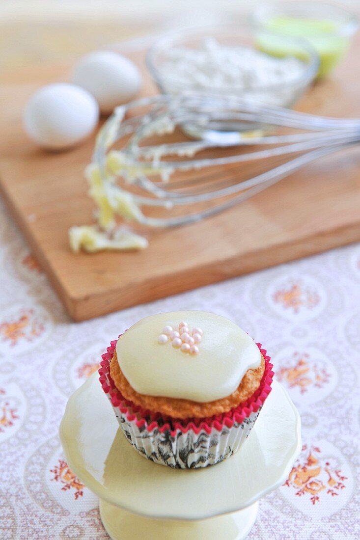 A muffin with icing and sugar balls with baking ingredients in the background