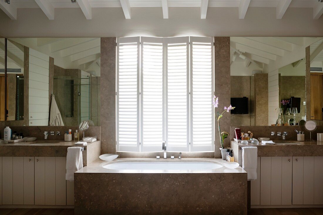 Marble bathtub below half-closed window shutters in spacious bathroom