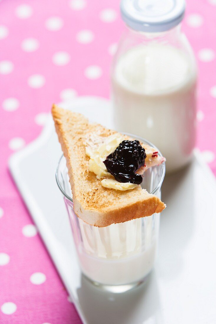 Blackberry jelly on a triangle of toast with a bite taken out of it on top of a glass of milk
