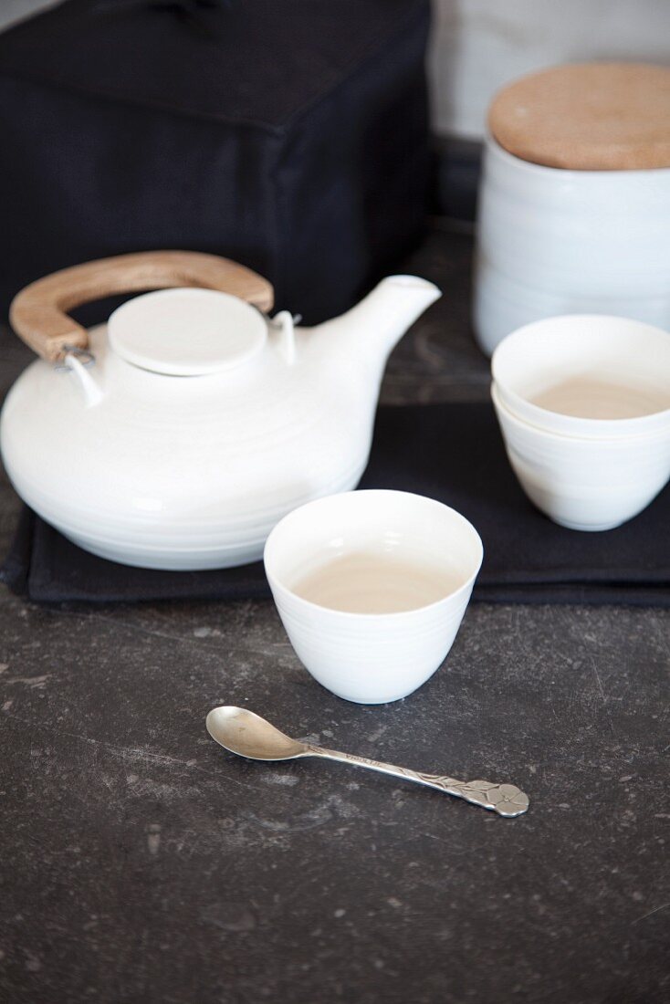 White tea set on grey stone surface; teapot with wooden handle, tea bowls and sugar box with wooden lid