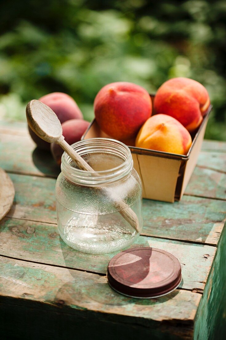 Offenes Schraubglas mit Holzlöffel und frische Pfirsiche auf einem kleinen rustikalen Tisch im Garten