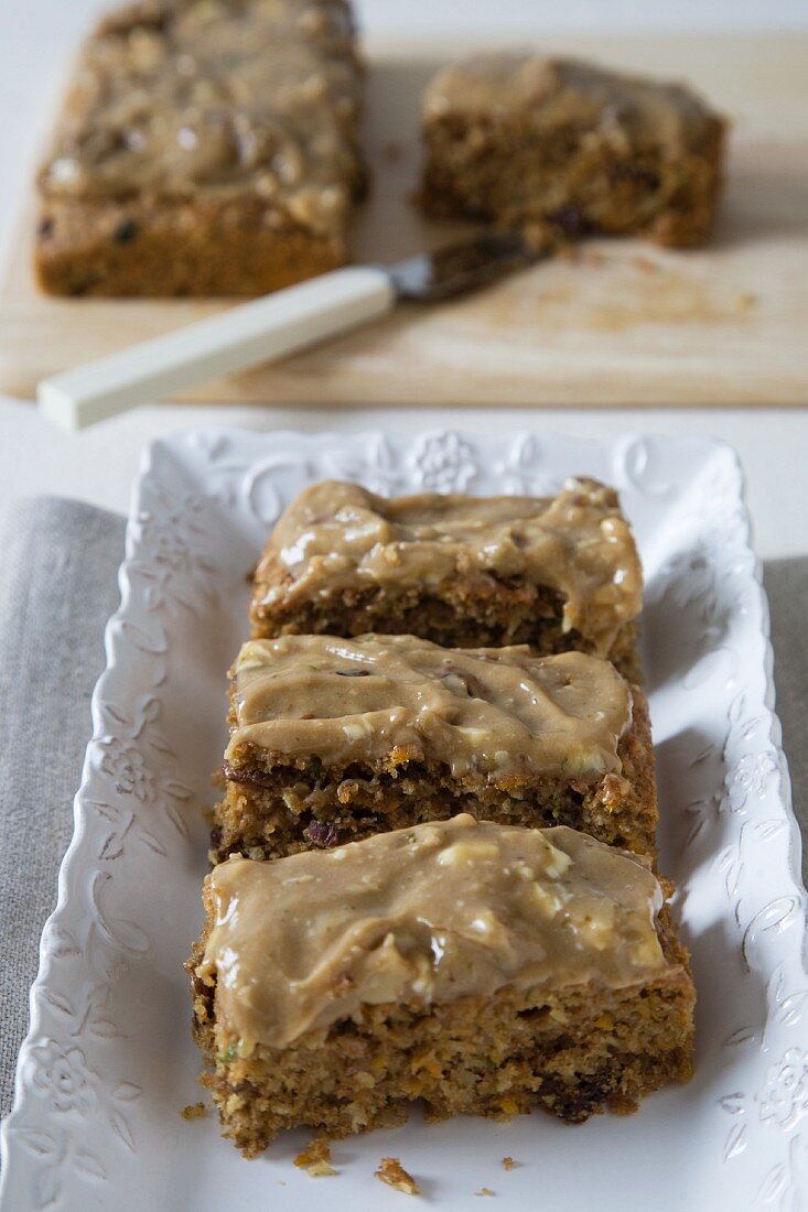 Three Pieces of Carrot Cake in a Rectangular Dish; Carrot Cake on a Cutting Board in the Background