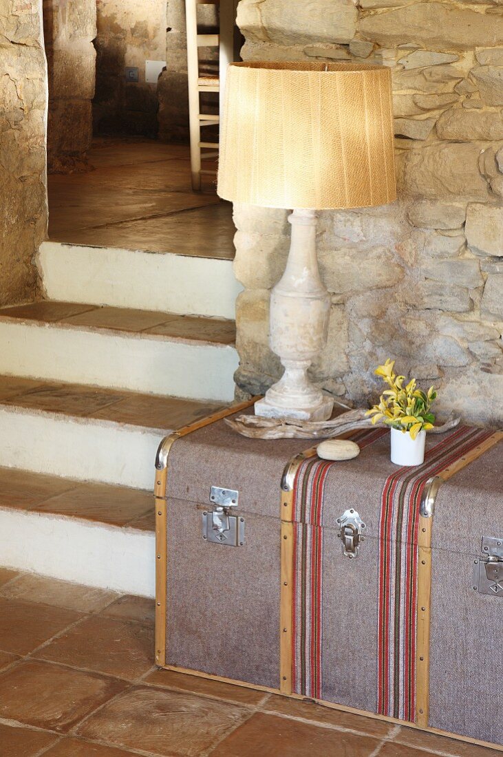 Traditional table lamp on steamer trunk against stone wall next to terracotta-tiled steps