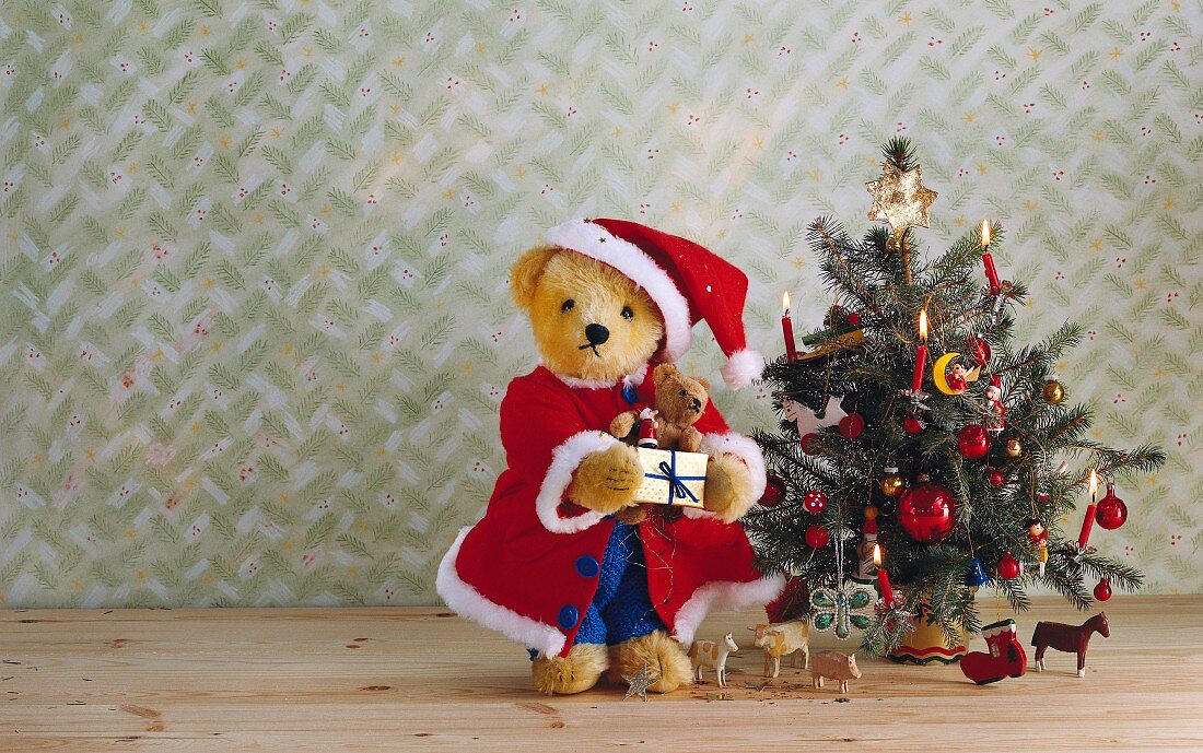 Teddy bear dressed as Father Christmas and toy ornaments next to Christmas tree