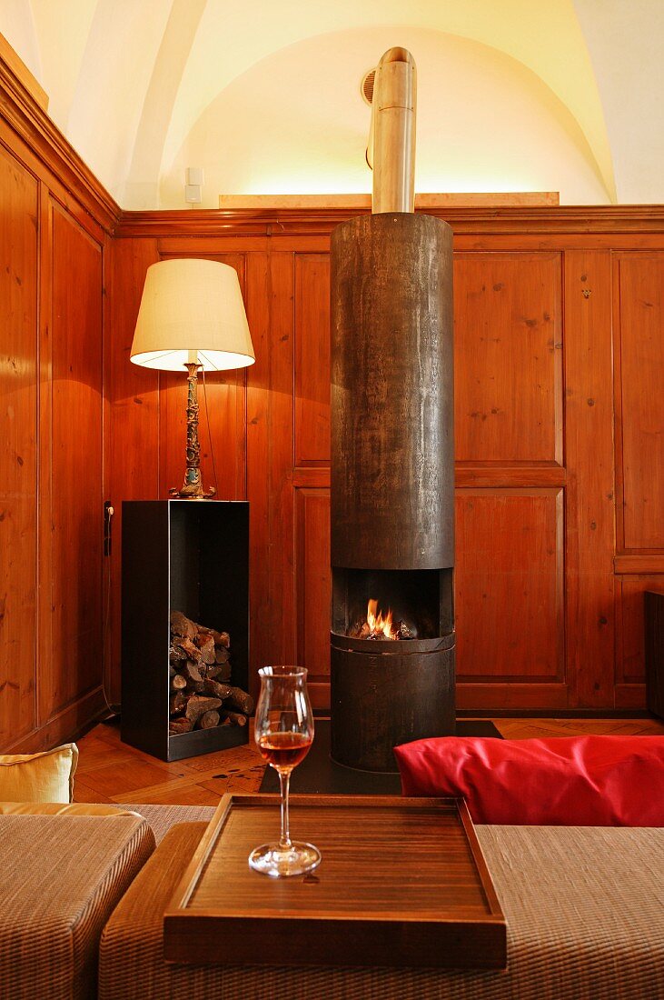 Antique-style table lamp next to modern, designer log burner made of unpolished steel in wood-panelled fireplace room of Schloss Schauenstein