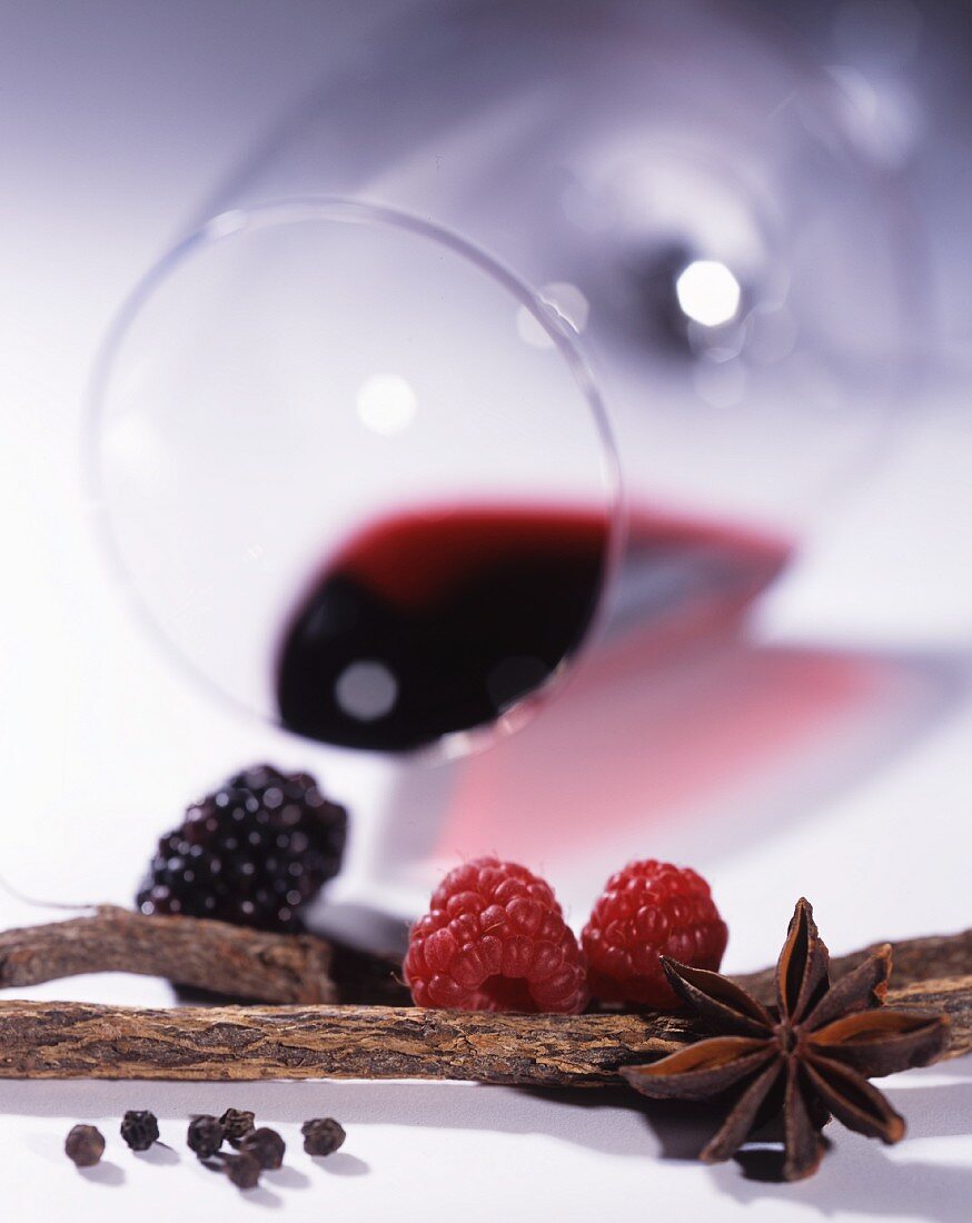 A glass of red wine lying on its side, with berries and spices