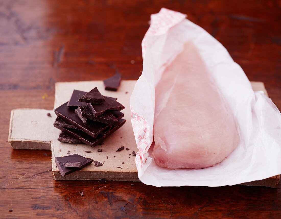 Raw chicken breast on a piece of paper and dark chocolate on a wooden board