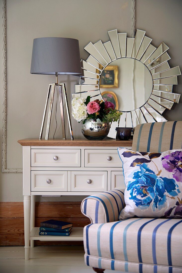 Lamp with lampshade and vase of flowers on cabinet in front of sun-shaped mirror