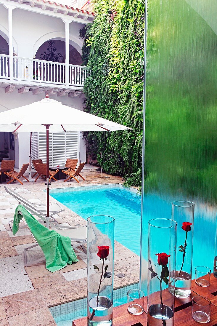 Red roses in glass vases on table and view down onto pool with loungers under parasols in colonial-style hotel courtyard