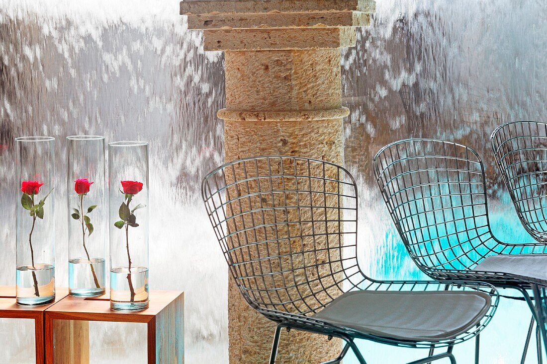 Stühle aus Drahtgestell und Rosen in Glasvasen auf Beistelltischen vor Steinsäule und Wasserfall in Pool stürzend