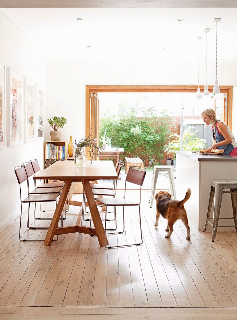 Delicate, chrome-framed chairs at wooden table opposite woman working at kitchen counter in modern interior with open terrace doors