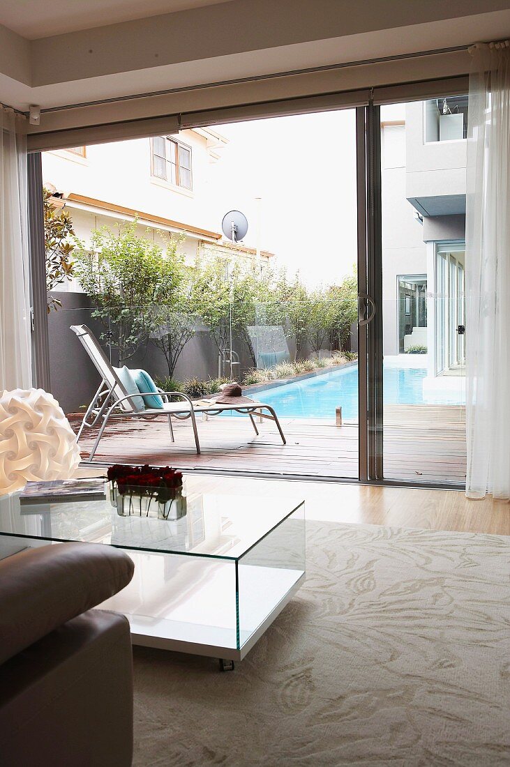 Kubischer Couchtisch aus Glas auf hellem Teppich vor offener Schiebetür und Blick über Terrasse auf Pool im Innenhof