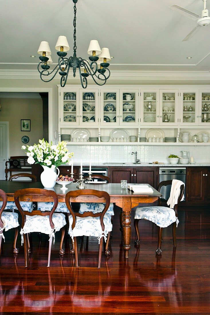 Antique style dining room with long dining table and romantic chair cushions on wooden chairs in an open living room with a stately, built-in kitchen