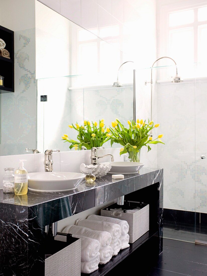 Bouquet of yellow tulips on marble washstand with towels on shelf and retro tap fittings; floor-level shower in background