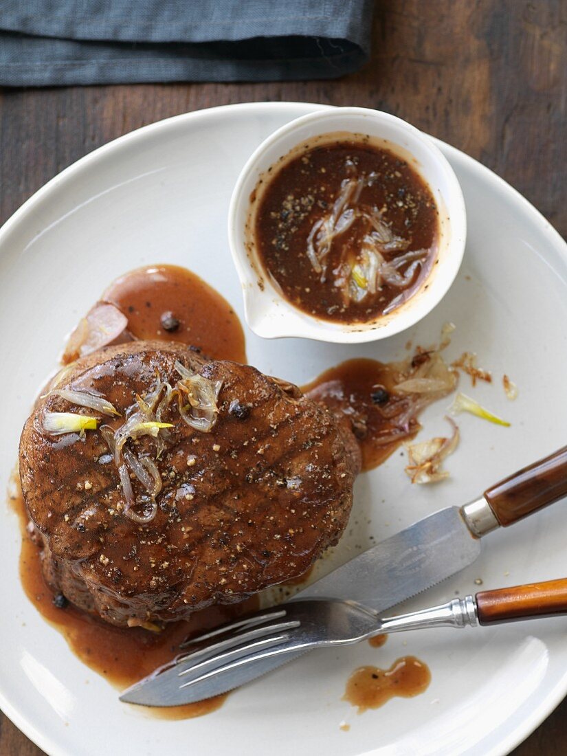Steak with Peppercorn Sauce and Shallots on a White Plate with a Fork and Knife