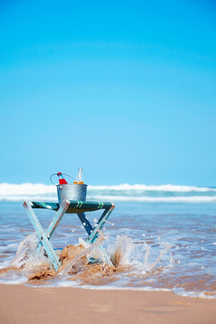 An ice bucket with drinks on a folding stool at the beach