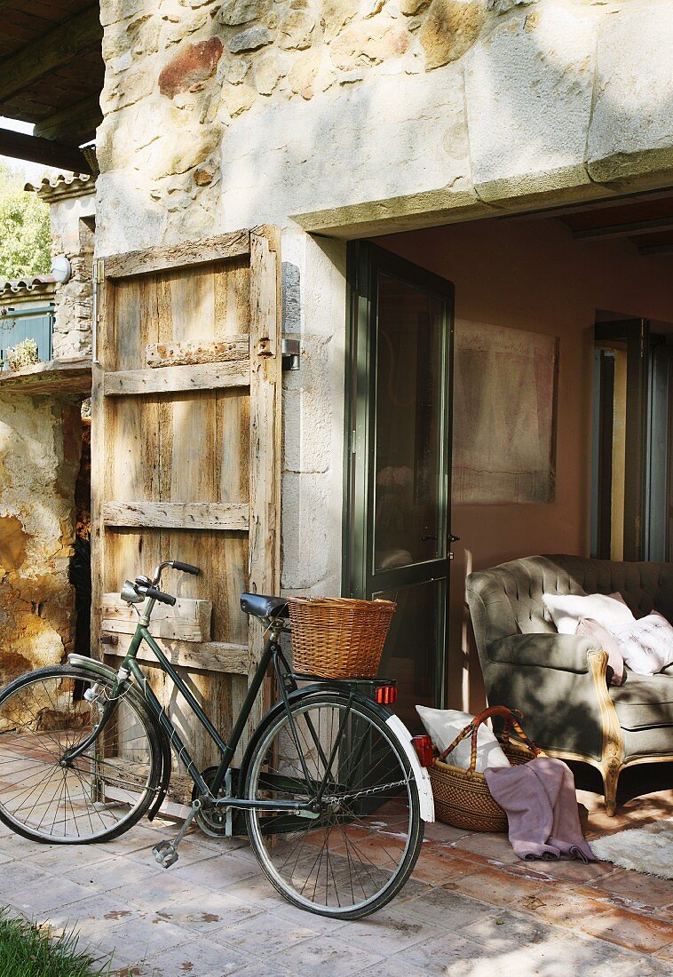 Bicycle with basket in front of stone house with door shutter and view of sofa through open terrace doors