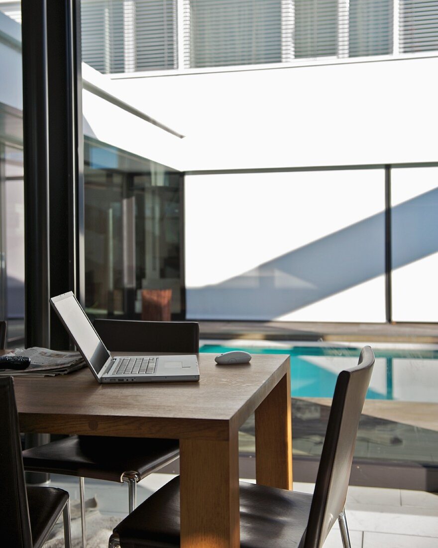 Open laptop on wooden table in front of glass wall with view of courtyard