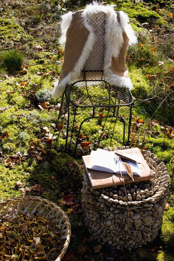 Felljacke über Drahtstuhl gehängt, davor ein kreatives Beistelltischchen mit aufgeschlagenem Notizbuch und Stift auf sonniger Waldlichtung