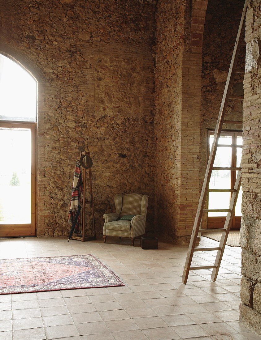 Wing-back armchair and coat rack in corner of high-ceilinged, restored, Mediterranean-style stone house