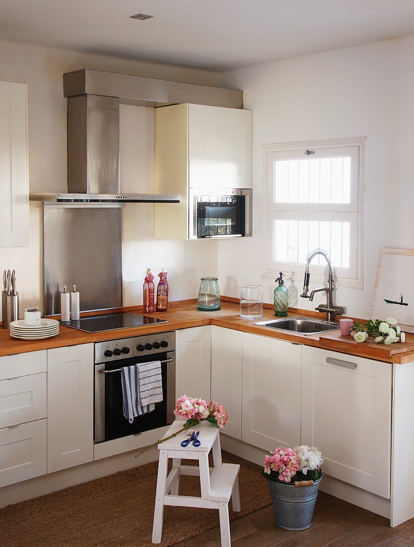 Kitchen corner with a window - flower bouquet in a pail and step stool in front of a kitchen counter with white lower cabinets and a stainless steel extractor fan above the stove