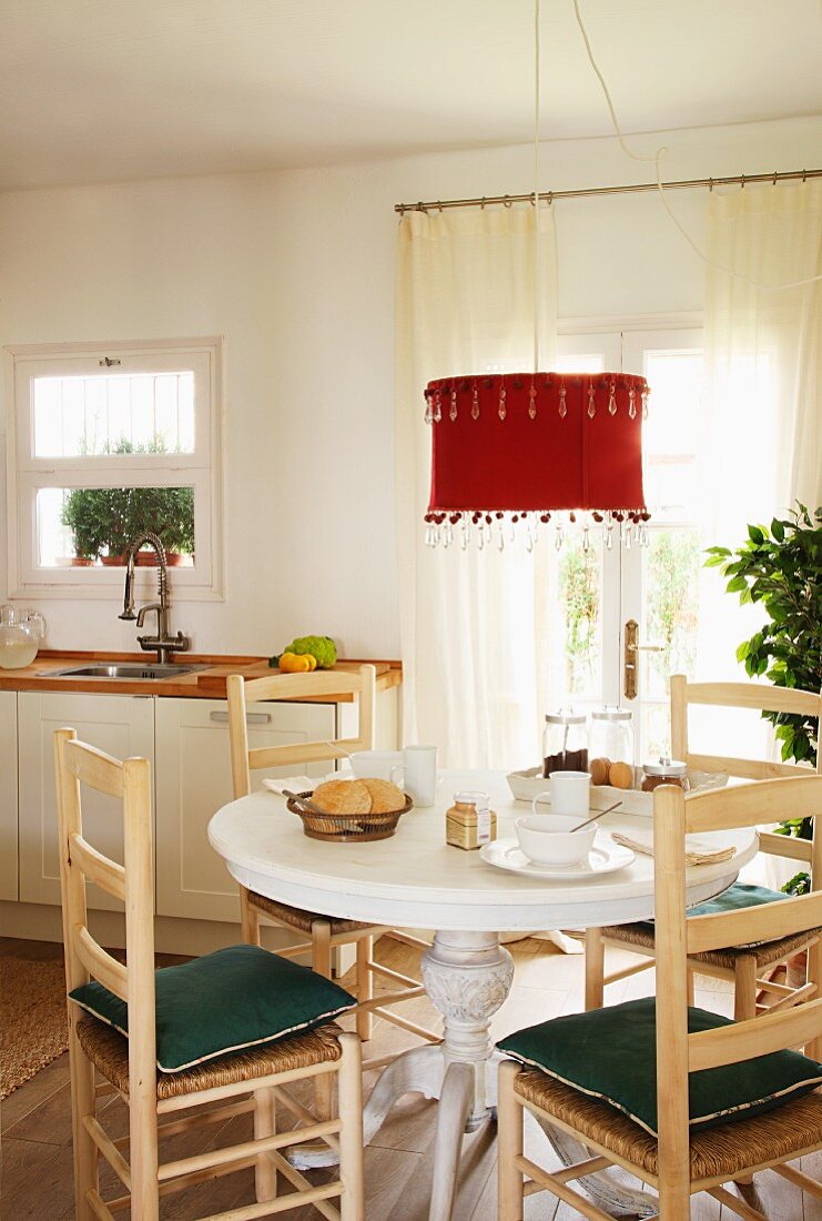 Mediterranean wooden chairs around a breakfast table under an hanging lamp with a red lampshade in front a of a patio door in a white kitchen