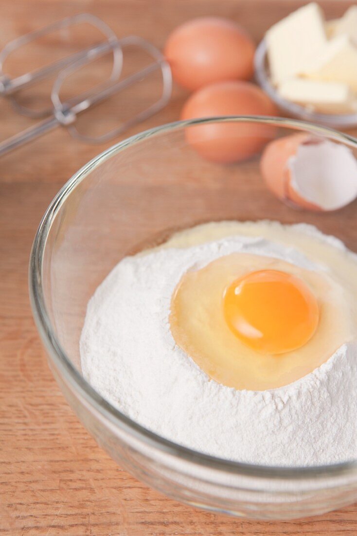A raw egg on top of flour in a glass bowl, alongside eggs, butter and whisks