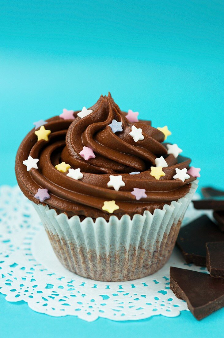A cupcake with chocolate icing and sugar stars in a paper case on a doily, and pieces of chocolate