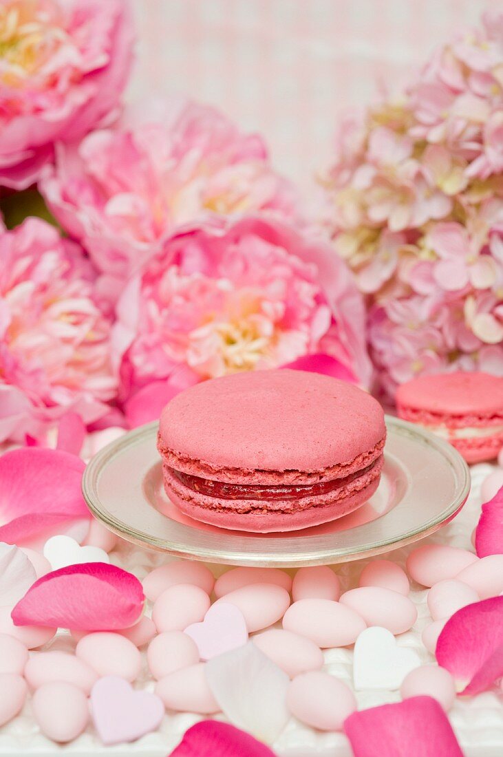 A pink macaroon on a silver plate, surrounded by sugared almonds, peonies and rose petals