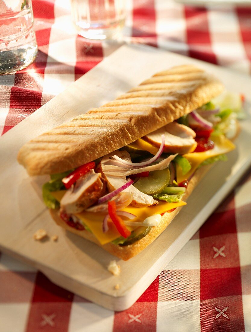 Baguettesandwich mit Hähnchen, Zwiebeln, Paprika, Essiggurken, Schmelzkäse, Salat und Ketchup auf Holzbrett