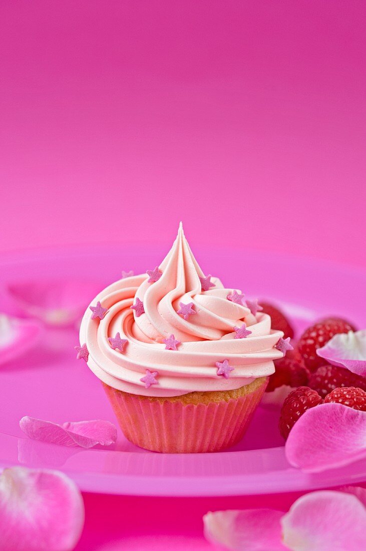 A pink strawberry cupcake with raspberries and rose petals