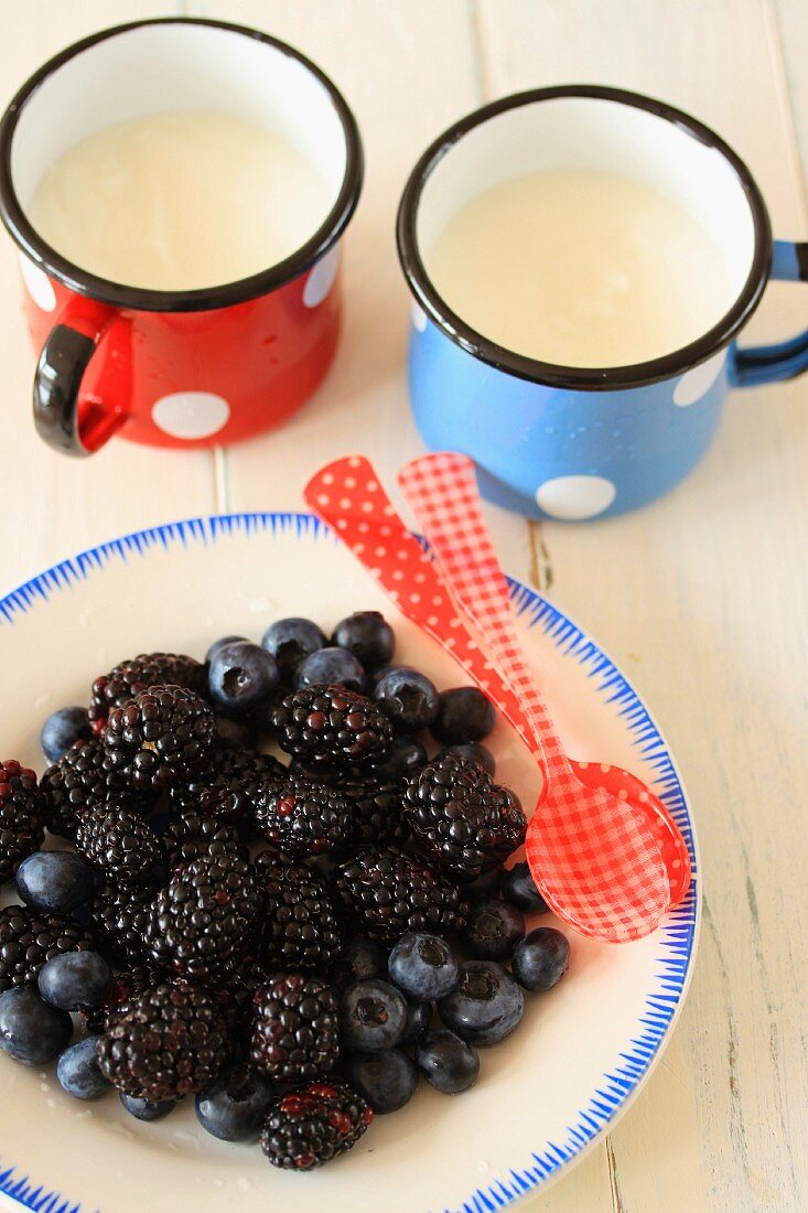 Blackberries and blueberries on a plate with two enamelled mugs of natural yoghurt