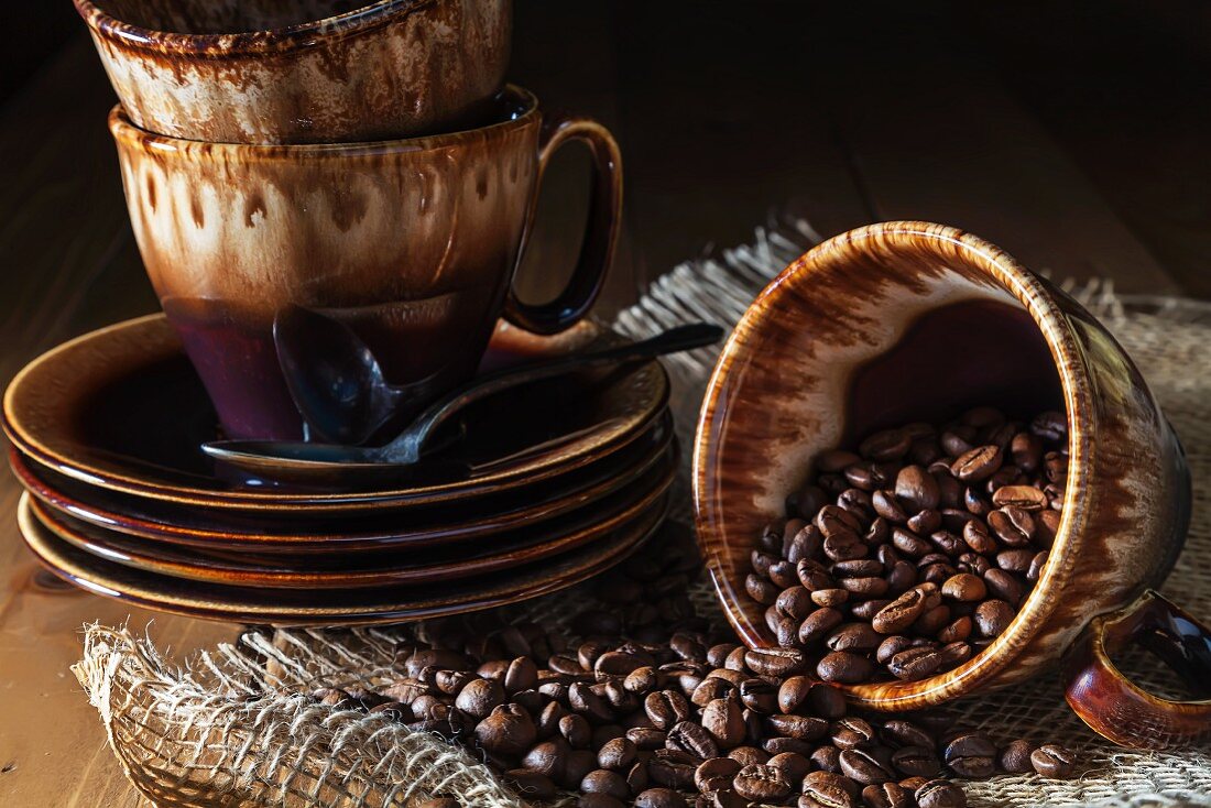Coffee Beans Spilling From a Tipped Cup on a Wood Table with Burlap; Stacked Plates and Cups