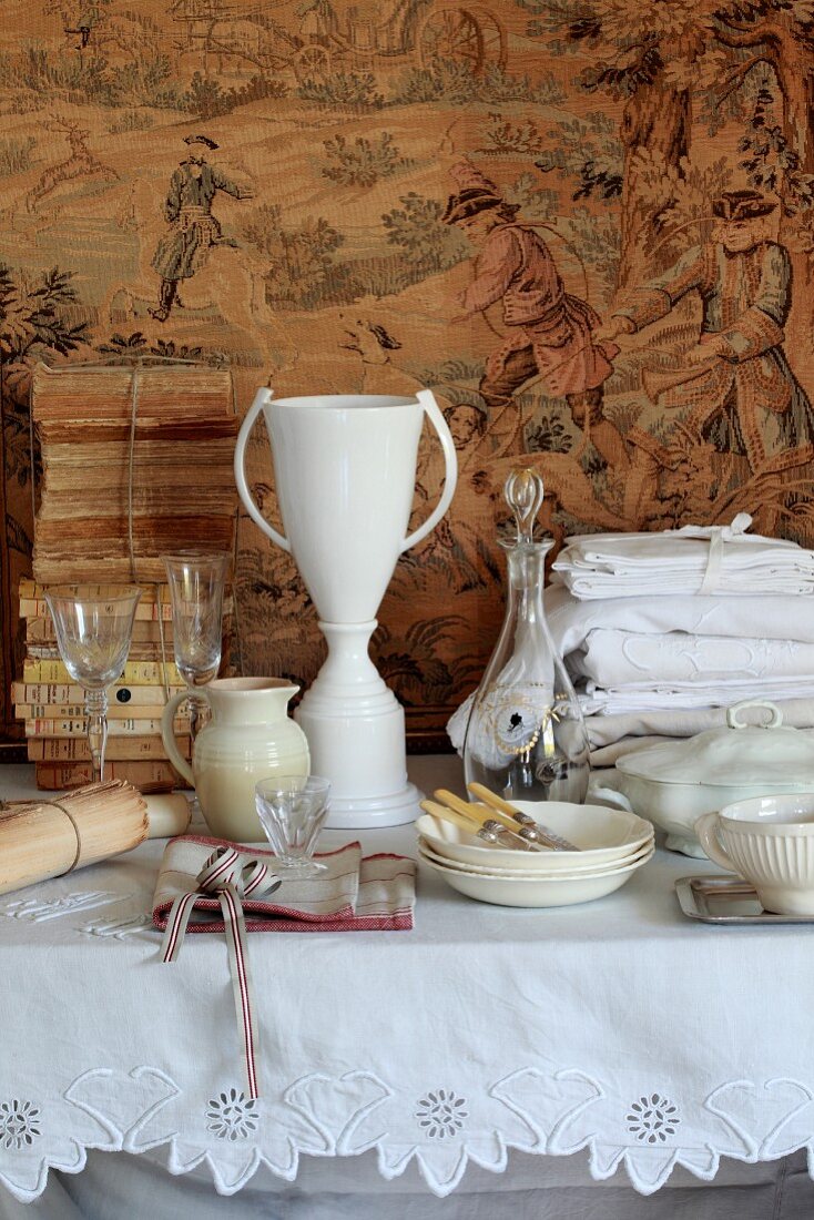 Glassware and crockery, vintage embroidered table linen and antique books on a table in front of a tapestry