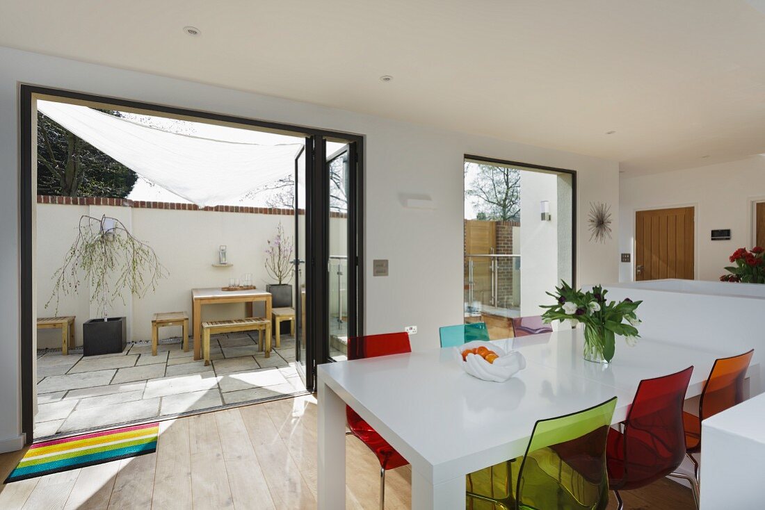 Farbige Kunststoff Stühle an weißem Esstisch vor offenen Terrassentüren und Blick auf Innenhof