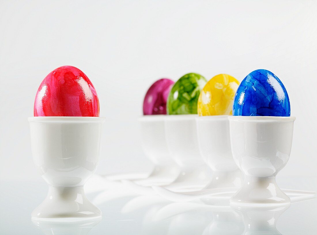 Five eggs, brightly coloured for Easter, in white eggcups with egg spoons