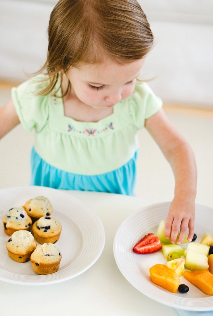 Kleines Mädchen mit klein geschnittenen Früchten und Blaubeermuffins