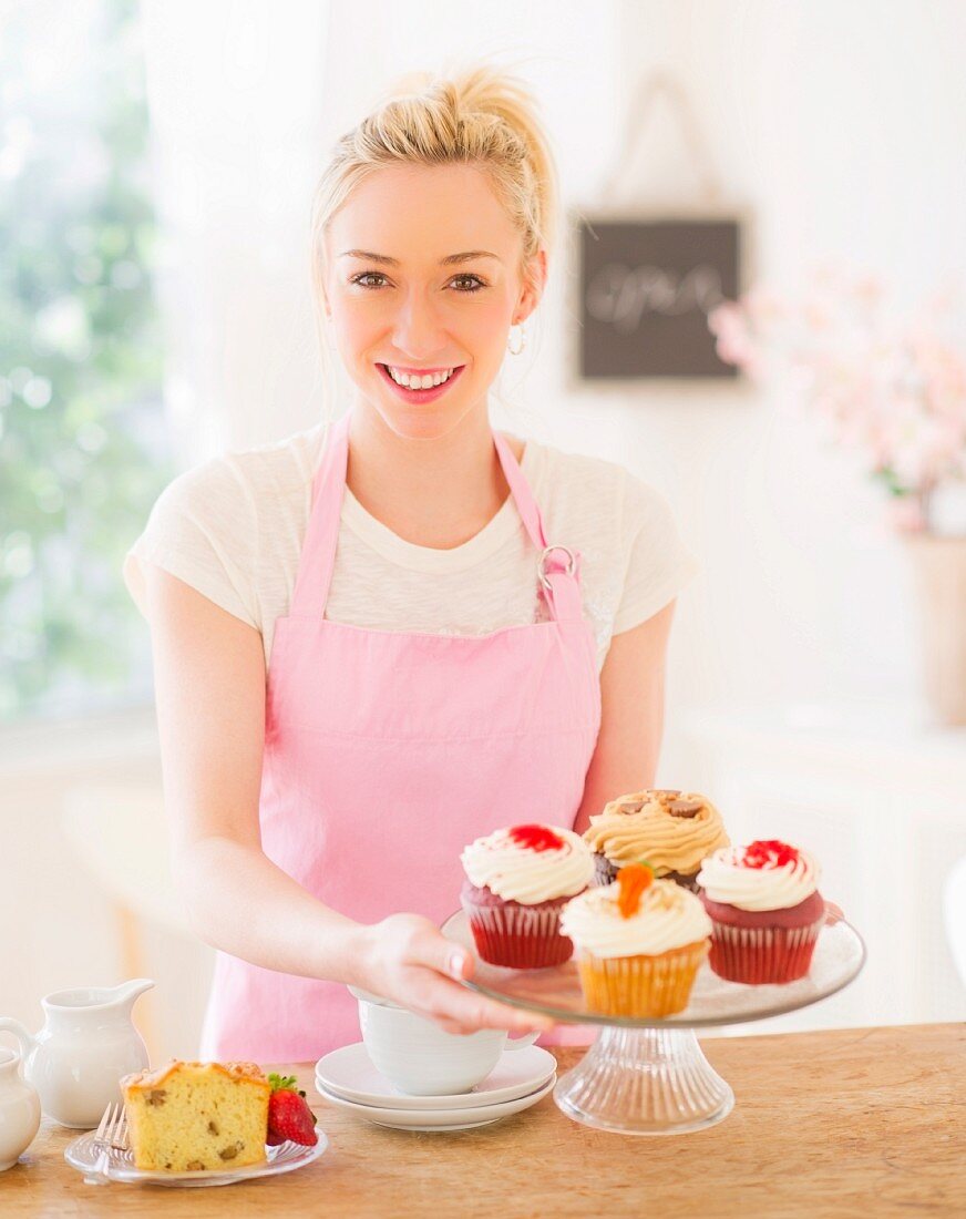 Lächelnde junge Frau mit Schürze hält Kuchenständer mit Cupcakes