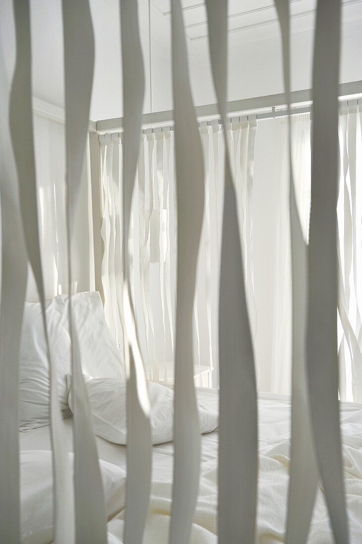 View through ribbons hanging from wooden frame of four-poster bed with white bedlinen