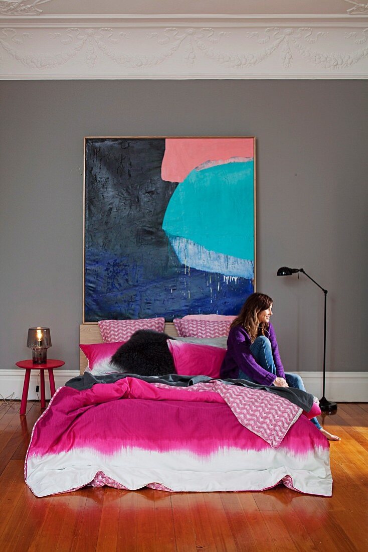 Doppelbett mit pinkfarbenem Bezug im Batiklook vor grossformatigem Farbgemälde; Frau sitzt auf Bett