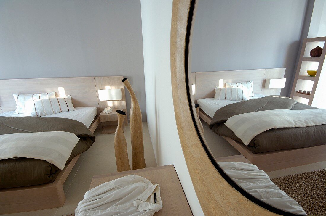 Round mirror on wall opposite modern double bed with pale wooden frame