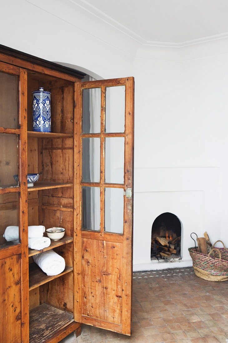 Old, glass-fronted cabinet with open door in interior with terracotta floor and open fireplace built into wall