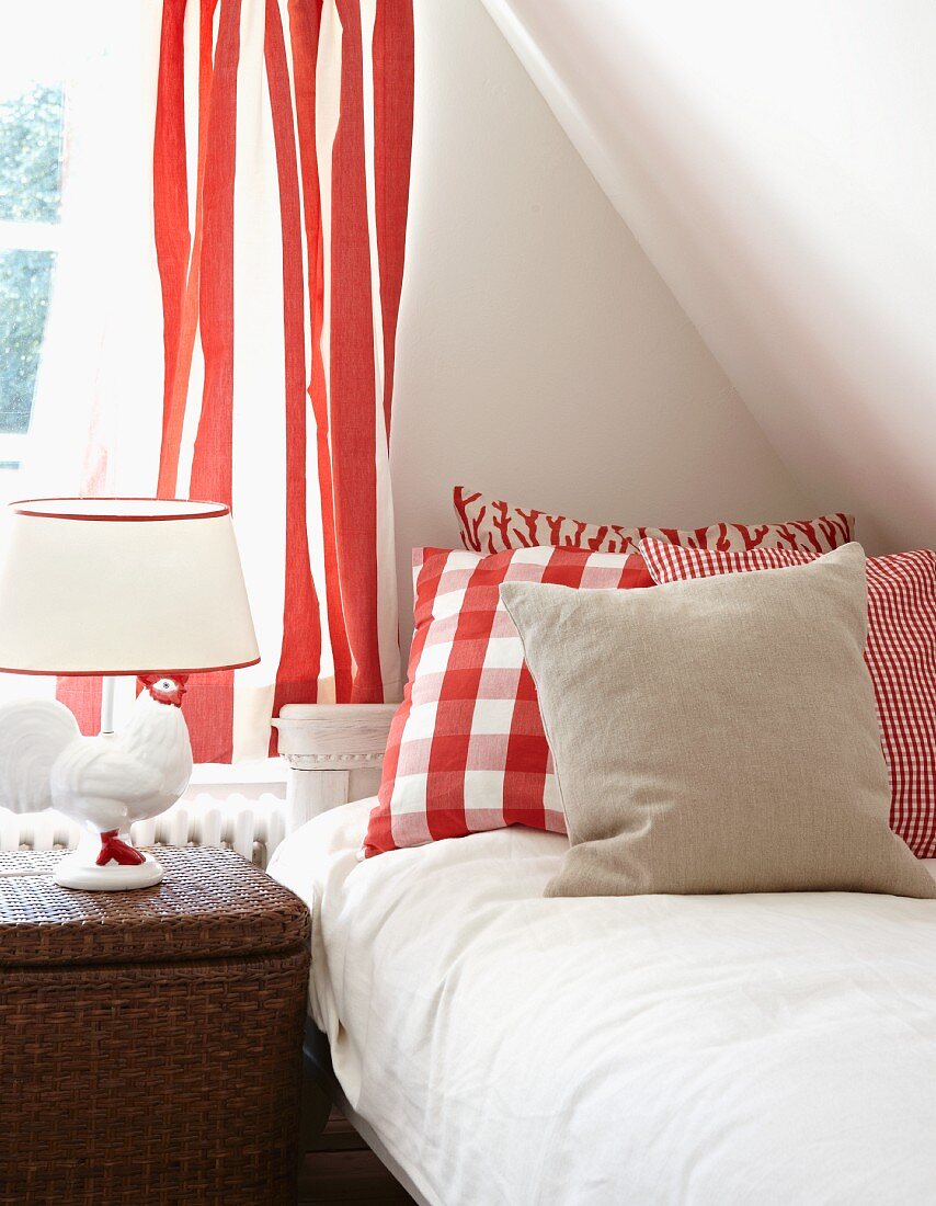 Bedside lamp with ceramic chicken base, red and white patterned curtain and scatter cushions at head of bed