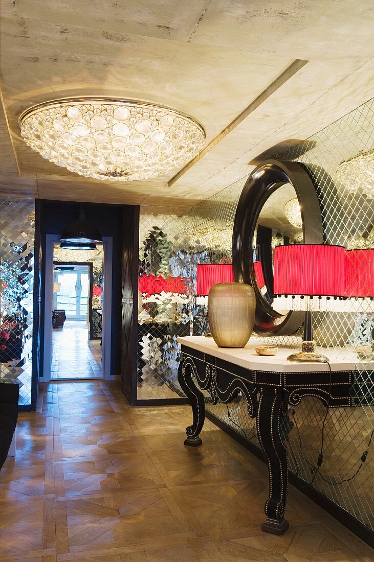 Console table with curved legs and table lamps with red lampshades against mirrored mosaic wall; elegant ceiling lamp on exposed concrete ceiling