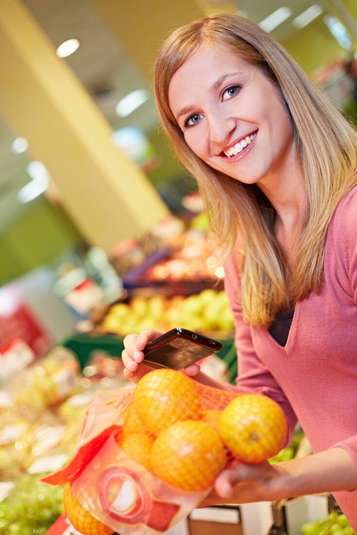 Germany, Cologne, Young woman with smart phone and oranges in supermarket, smiling