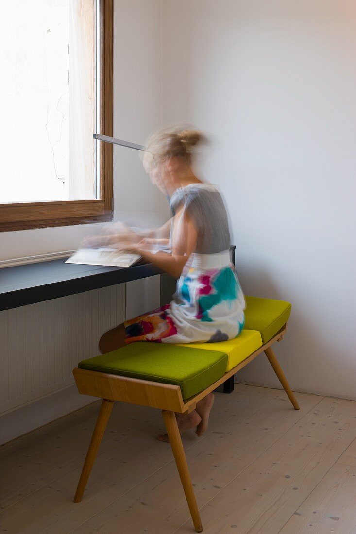 Lady sitting on a bench with a green pad at a minimalist table in front of a window