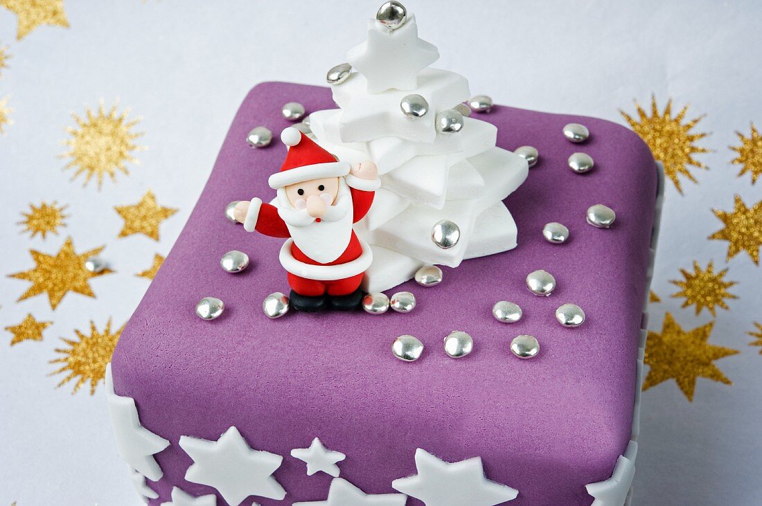 a model in icing of father Christmas holding up his arms on a purple and white Christmas cake with white stars and golden stars on a white background.