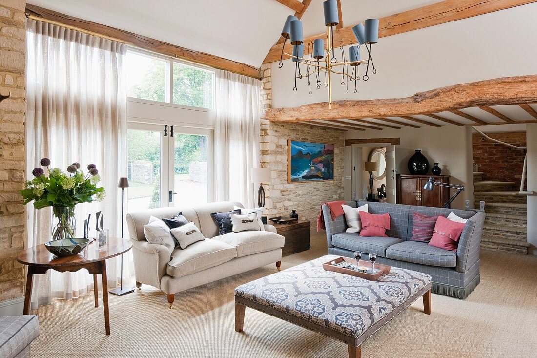 Elegant living room with rustic stone walls and wooden beams; two vintage-style sofas and square ottoman in centre of room