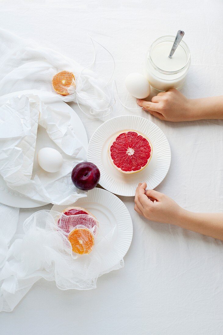 Hands on a table with grapefruit, a plum, eggs, white plates and a muslin cloth